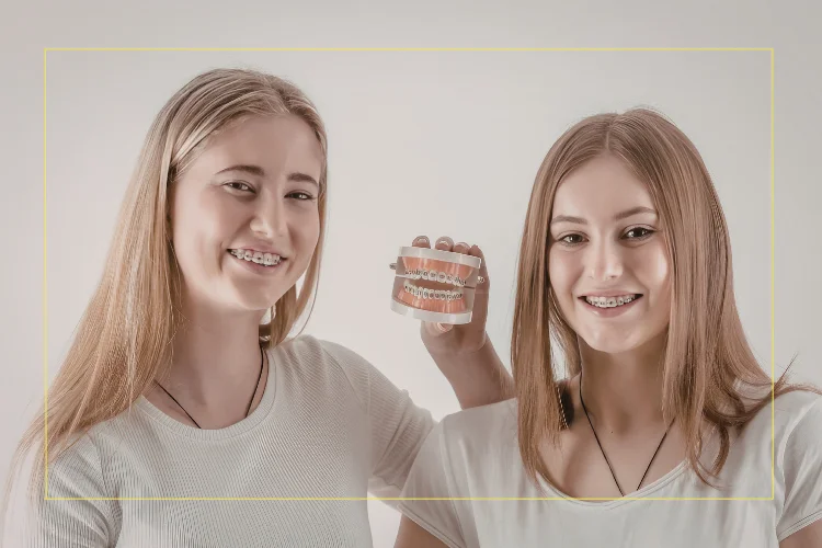 Two smiling teenage girls wearing braces, with one holding a dental model showing orthodontic brackets and wires